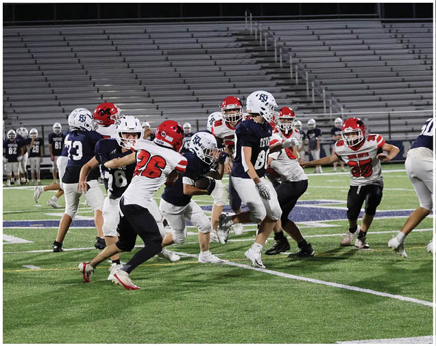 A group of people wearing football uniforms with red and white or navy and white helmets are actively playing on a green field with empty bleachers in the background. Description generated by AI