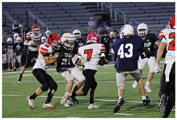 Three people wearing football uniforms are engaged in a play on a grassy field, with two in white jerseys and one in a dark jersey. Description generated by AI