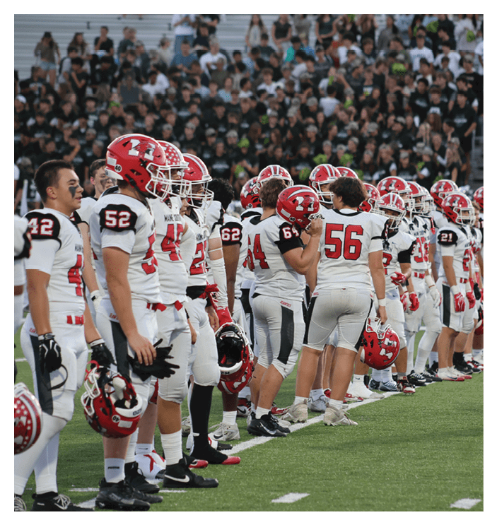 A line of people in white football uniforms with red helmets stand on a field facing a crowd in the background. Description generated by AI