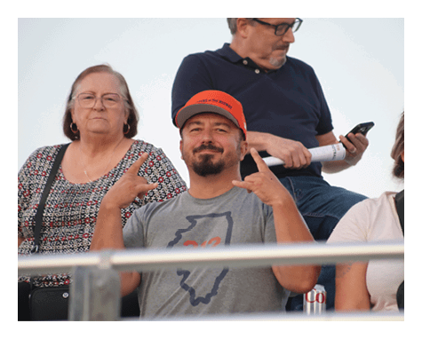 A person wearing a gray t-shirt and an orange cap is smiling and making hand gestures while sitting between two other people, with another person behind them looking at a phone. Description generated by AI