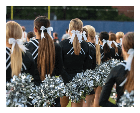 A line of people dressed in black cheerleading uniforms with white bows in their hair hold silver pom-poms while standing outdoors on a field. Description generated by AI