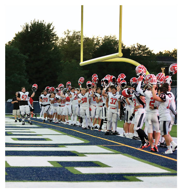 A group of people in football uniforms stand on the sideline of a football field near the end zone, many holding their helmets up in the air. Description generated by AI