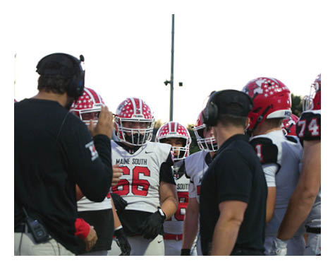 A group of football players in white and red uniforms are gathered closely with two coaches wearing headsets during a game or practice. Description generated by AI