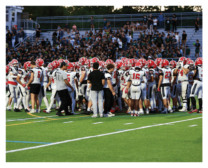 A large group of people wearing white and red football uniforms and helmets are gathered on a green field with some people in casual clothing standing among them. Description generated by AI