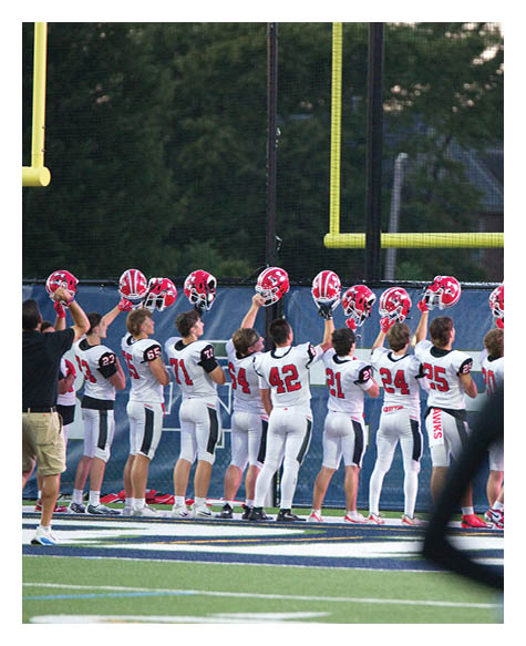 A group of people in white sports uniforms stand in a line on a football field holding red helmets up toward the sky near a goalpost. Description generated by AI