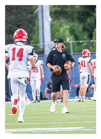 A person wearing a black outfit and cap is holding two footballs on a field while several people in white and red football uniforms stand in the background. Description generated by AI
