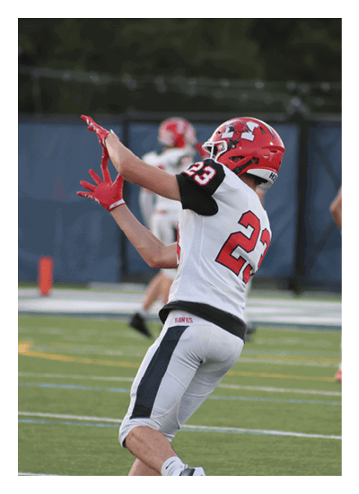 A person wearing a red helmet and white football uniform with the number 23 is reaching up to catch a football on a green field with other players in the background. Description generated by AI
