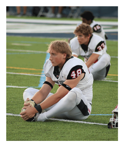 A person in a white football uniform with the number 48 is sitting on the grass field stretching their legs while two other people in similar uniforms are sitting behind them. Description generated by AI