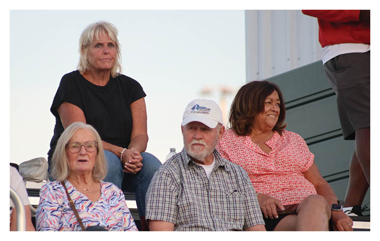 Three adults are sitting closely together on outdoor bleachers, with one wearing a white cap and plaid shirt, another in a black shirt, and the third in a patterned blouse and glasses. Description generated by AI