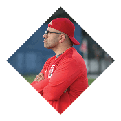 A person wearing a red cap, glasses, and a red long-sleeve shirt is standing with arms crossed, looking intently to the side in an outdoor setting. Description generated by AI