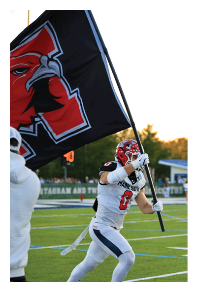 A person in a white football uniform and helmet is running on a field while holding a large black flag with a red and white design. Description generated by AI