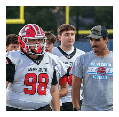 A person wearing a red football helmet and a white jersey with the number 98 and the words Maine South stands on a football field near a person in a black shirt and gray pants. Description generated by AI