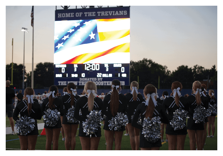 A group of cheerleaders with silver pom-poms and white hair ribbons stand on a field facing a large scoreboard displaying an American flag and game information at dusk. Description generated by AI
