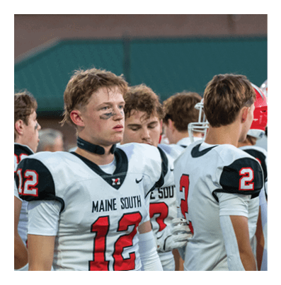 A group of people wearing white and black football uniforms with red numbers stand together outdoors, with one person in the foreground wearing number 12 and black face paint under their eyes. Description generated by AI