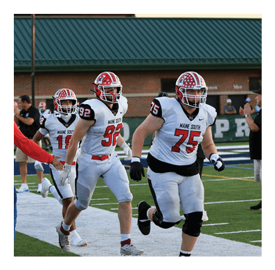 Several people in football uniforms are running onto a field from the sidelines while a person in a red jacket and cap reaches out to them. Description generated by AI