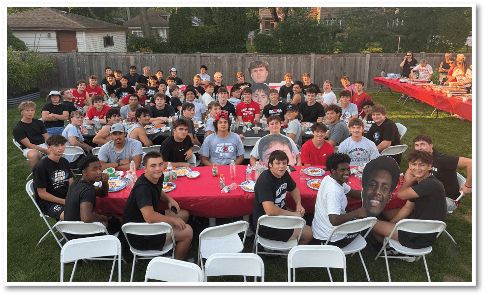 A large group of people sitting around tables with red tablecloths outdoors on grass, enjoying a meal together in a backyard setting with trees and houses in the background. Description generated by AI