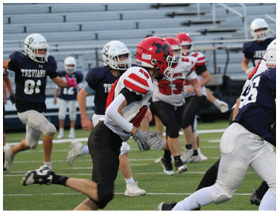 Children wearing football helmets and uniforms are engaged in a game on a field with empty bleachers in the background. Description generated by AI