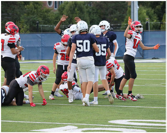 People wearing football helmets and uniforms are gathered on a field with some standing and others sitting or kneeling, appearing to be in the middle of a game or practice. Description generated by AI
