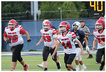 Two people wearing football uniforms are running on a field, one in a white and red jersey with the text “MAINE SOUTH" and the other in a navy blue jersey. Description generated by AI