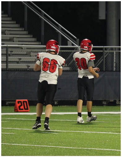 Four people wearing football helmets and uniforms stand on a field with one person in a white and red uniform facing away from the camera. Description generated by AI