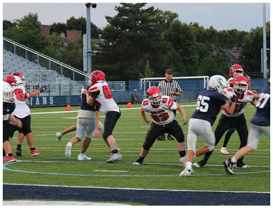 Children wearing football helmets and uniforms are engaged in a game on a field with empty bleachers in the background. Description generated by AI