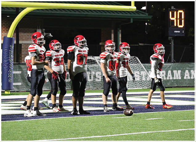 Several people wearing red and white football uniforms stand on a green field near a goalpost and a football resting on the ground. Description generated by AI
