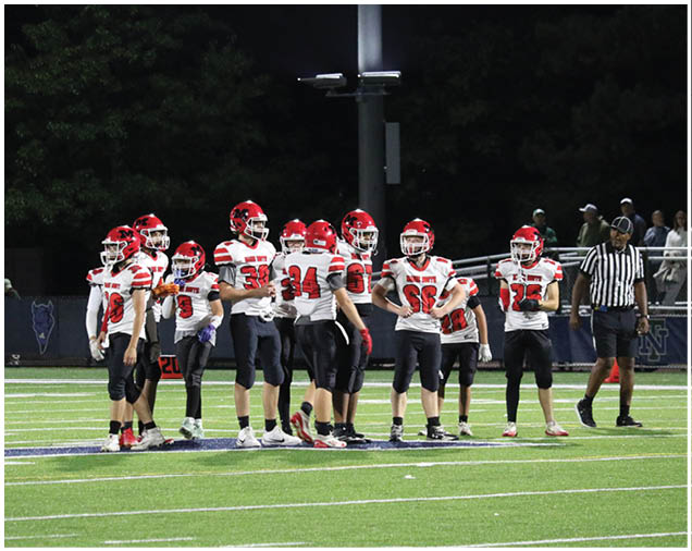 A group of people wearing red helmets and white sports jerseys stand together on a football field at night with a referee nearby. Description generated by AI