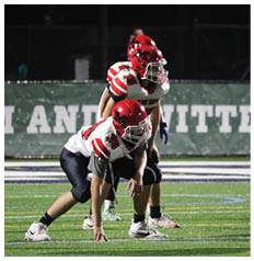 A person in a red helmet and white football jersey with red stripes is crouched on a football field, preparing for a play. Description generated by AI