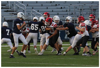 People wearing football uniforms are actively playing on a field with one person in a red helmet preparing to catch or block while others in dark jerseys move toward them. Description generated by AI