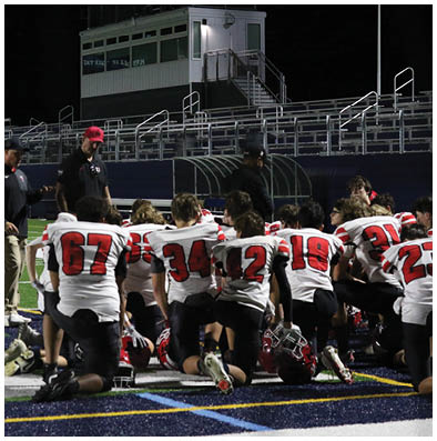 People wearing football uniforms with red numbers are kneeling on a field, facing away from the camera, with coaches standing nearby. Description generated by AI