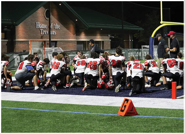 A group of people wearing football uniforms are kneeling and sitting on a field near a sideline marker during a nighttime game. Description generated by AI