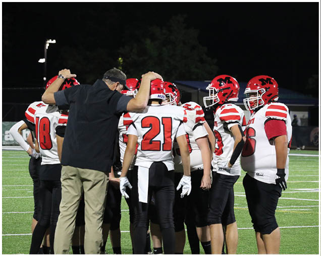 A group of football players in red helmets and white jerseys are huddled on a field at night with a coach giving instructions. Description generated by AI