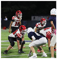 Two people in football uniforms and helmets are crouched facing each other on a field, preparing for a play. Description generated by AI