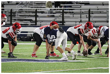 Three people wearing red helmets and white football jerseys are positioned on a football field, preparing for a play. Description generated by AI