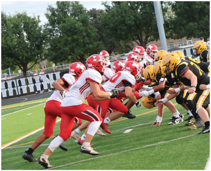 Two opposing American football teams in red and yellow uniforms are lined up facing each other on a green field, ready for the next play. Description generated by AI