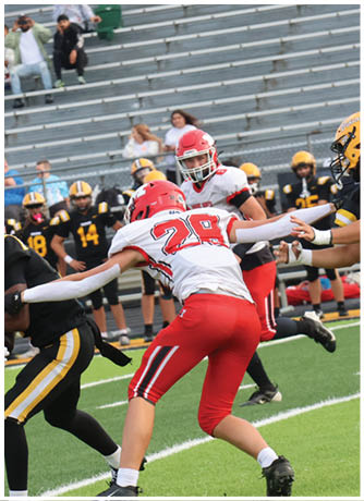 Two people in red and white football uniforms are actively playing against opponents in black and yellow uniforms on a green field with empty bleachers in the background. Description generated by AI