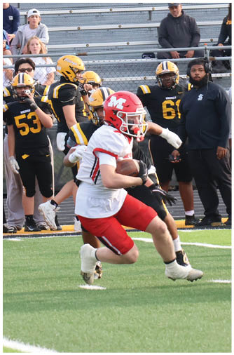 Players in football uniforms are actively engaged in a game on a green field with spectators and coaches watching from the sidelines and bleachers. Description generated by AI