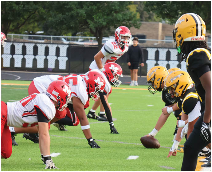 Two opposing American football teams in red and yellow helmets are lined up on the field preparing for a play. Description generated by AI