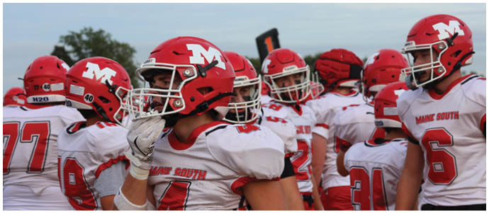 A group of people wearing red helmets and white sports uniforms with “Maine South" written on them are gathered together, likely preparing for or participating in a football game. Description generated by AI