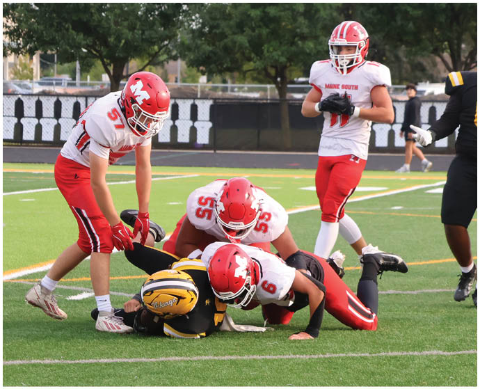 Three people in red and white football uniforms are tackling a person in a black and yellow football uniform on a green field while a referee and another player watch. Description generated by AI