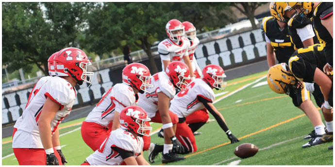 Two opposing football teams in red and black uniforms are lined up facing each other on a field, ready for a play. Description generated by AI