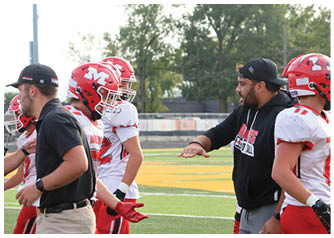 A coach wearing a black hoodie and cap is giving instructions to football players in red and white uniforms on a field. Description generated by AI
