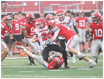 Several people wearing football helmets and uniforms are actively engaged in a tackle on a grassy field with spectators in the background. Description generated by AI