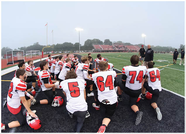 A group of people wearing white and red sports uniforms are kneeling on a football field listening to two standing coaches during a foggy day. Description generated by AI