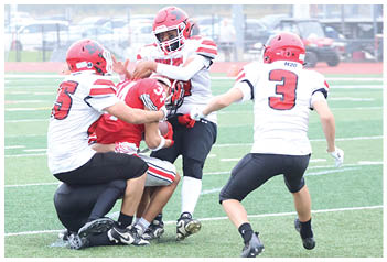 Four people wearing football helmets and uniforms are engaged in a tackle on a green football field. Description generated by AI