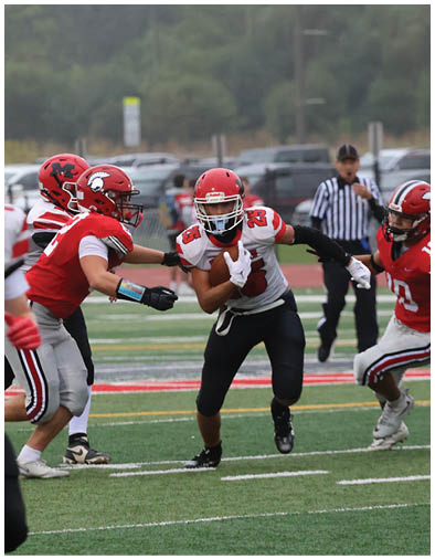 A person in a white and black football uniform is running with the ball while three people in red and white uniforms attempt to block or tackle on a football field. Description generated by AI