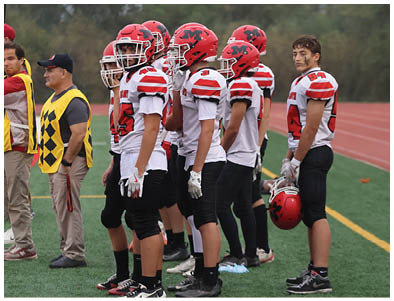 A group of people wearing red and white football uniforms stand on a green field near a running track with two adults in yellow vests nearby. Description generated by AI