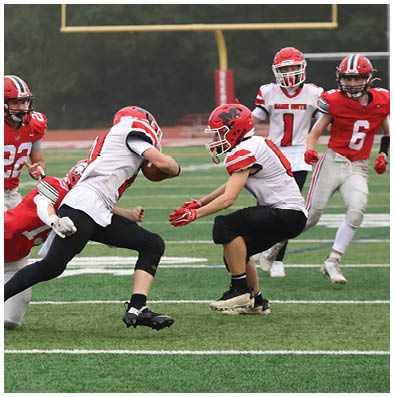 Two people in football uniforms with red helmets are engaged in a play on a football field while two others in similar uniforms watch nearby. Description generated by AI