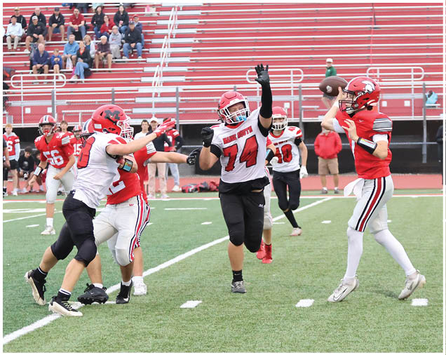 A group of people wearing football uniforms are playing on a field with one person in red preparing to throw a football while others in white and red attempt to block or tackle. Description generated by AI