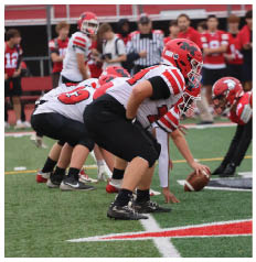 Several people in football uniforms are lined up on a field preparing for a play, with one person holding a football ready to snap it. Description generated by AI